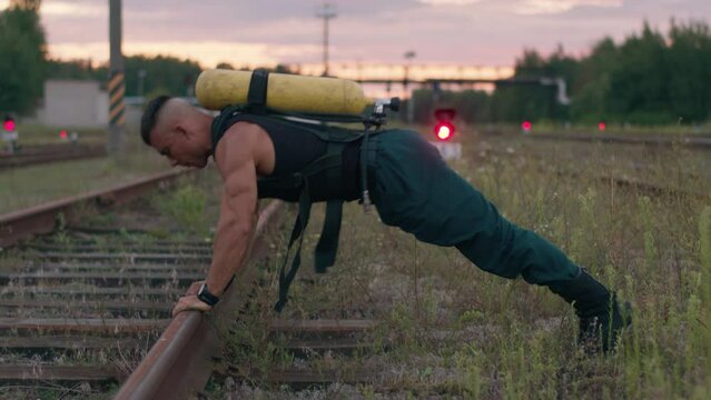 Young muscular man training on the railway tracks and doing push-ups from the rails with a balloon drager on his back