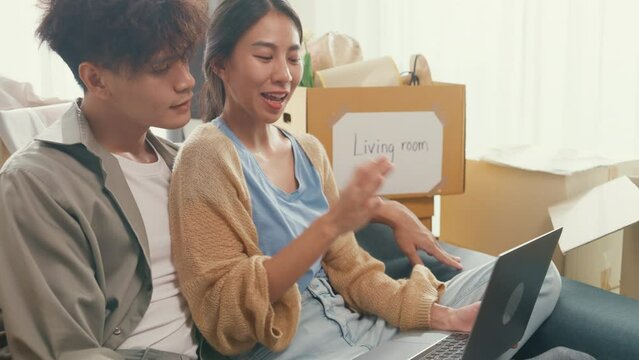 Young happy Asia couple sit on sofa couch use computer laptop online shopping furniture from decoration store in living room at home. Husband wife move new house, Apartment Rent And Ownership Concept.