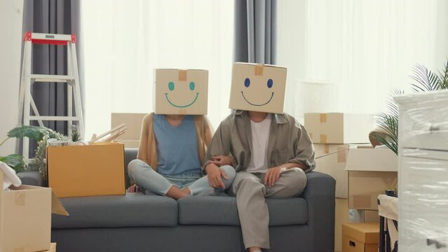 Happy Asian couple sit on sofa couch used carton box made smiling face funny dance front of camera in living room at new house. Husband and wife moving new house, Apartment Rent And Ownership Concept.