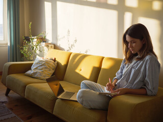Woman with laptop and notebook sitting on the sofa at home