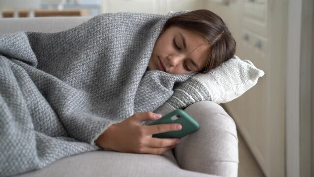 Unhealthy teenage girl coughing lying on couch after being infected with flu virus due to sharp cold snap and low temperature at school. Sick sad European child wrapped in blanket uses mobile phone