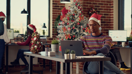 Diverse team of colleagues with santa hats working on laptop during winter holiday season in festive decorated office. Coworkers doing teamwork on computer, celebrating christmas eve.