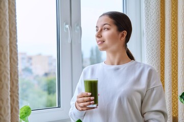 Teenage girl with glass of green fresh in morning at home near window