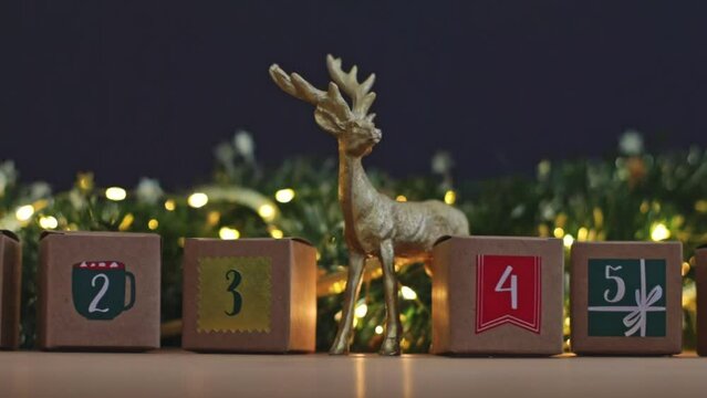 Beautiful advent calendar three kraft boxes with numbers 2,3,4,5 with deer christmas decor and a burning garland on a wooden table, bottom view close-up in slow motion and zoom out.