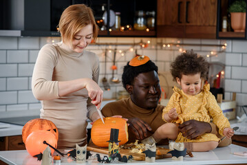 Fototapeta premium Multi-ethnic family with small child carving a pumpkin preparing it for Halloween holiday, making jack-o-lantern