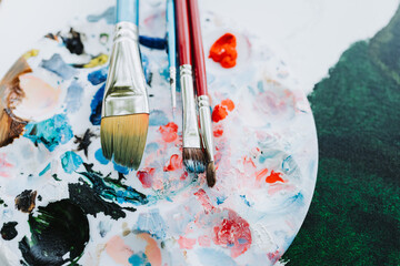 Close up of a round paint palette and brushes over a canva painted with green oil. Amateur painter.