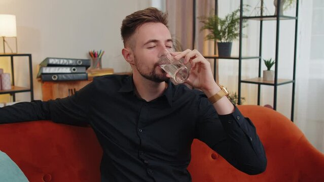 Portrait of thirsty lovely man sitting indoors holding glass of natural aqua make sips drinking still water preventing dehydration. Guy with good life habits, healthy slimming, weight loss concept