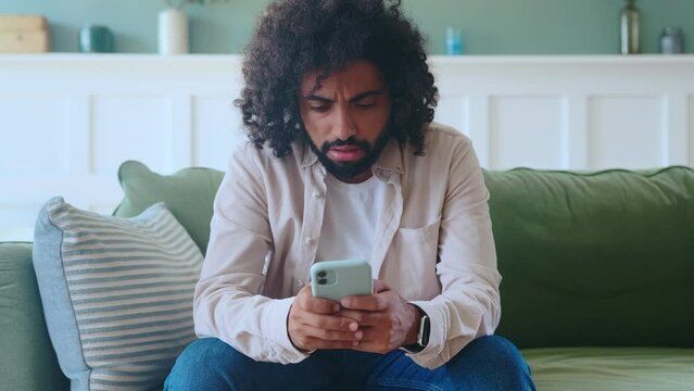 Young disgruntled Arabian man with phone in hand, chats with support service in mobile application or reads email and nervously drops smartphone sits on sofa in living room. Bad news concept