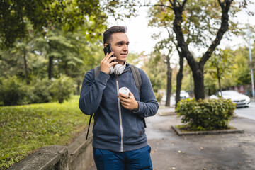 One young adult caucasian man walking in the city town near park using mobile phone smartphone talk with cup of coffee in autumn or spring day happy male tourist standing alone real people copy space