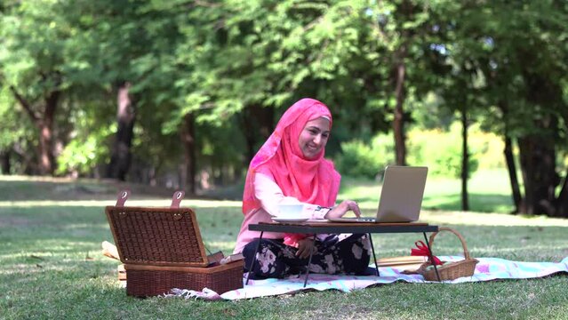 Asian muslim girl with hijab sit in park with picnic basket dring coffee talking with friends by using laptop, relaxing freelancer lifestyle working from everywhere with basket and coffee cup
