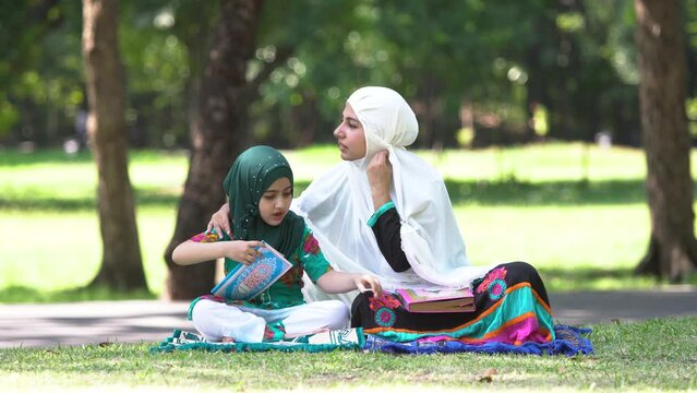 Muslim mother teaching her daughter to pray Quran bible, happy mom with hijab and cute daughter sitting in park spent holiday together, muslim family lifestyle motherhood relationship concept
