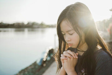 Preteen mixed girl praying with eyes closed by lake, kid worshiping online at home, peace and hope...