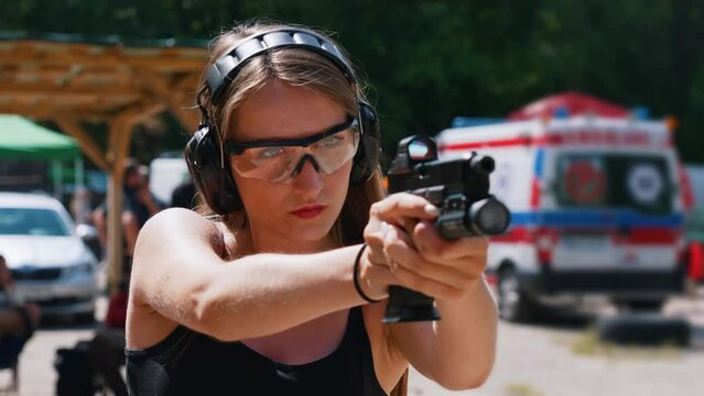 Powerful focused caucasian long-haired girl in protective gear aiming via loupe. Shooting range day. Outdoor medium closeup shot. High quality 4k footage