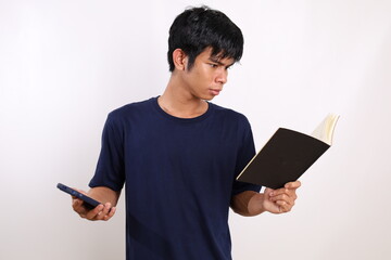 Serious expression of young asian man while reading a book and holding a cell phone. Isolated on white