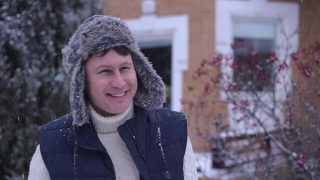 Joyful happy Caucasian man in fur hat posing in slow motion on snowy backyard and looking around admiring snow falling. Portrait of relaxed cheerful guy looking at camera smiling