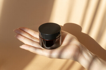 Amber jar with cream on woman's palm, on brown background in morning rays of sun. Concept of mockups, skin care