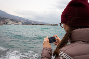 A woman takes pictures on her phone of waves in the sea on the shore of Yalta. Front view.