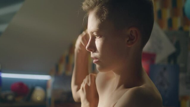 Focused boy looks at his biceps in front of the mirror in his room after exercise. Shirtless boy checks workout progress and training results. Athelete young boy. Memeber of school sports team.