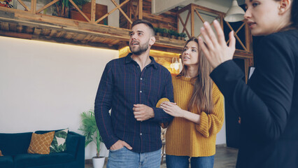 Confident housing agent is showing spacious modern house with beautiful interior to happy young couple wife and husband, people are smiling and talking looking around.