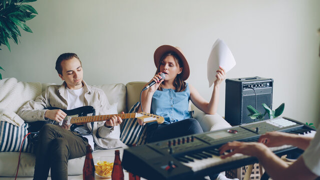 Cheerful young people musical group are practising in home studio singing and playing the keyboard and the guitar. Musicians are wearing trendy casual clothes.