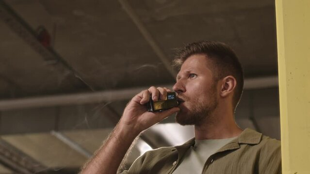 Low angle chest up slowmo of pensive young Caucasian man vaping on dark parking lot
