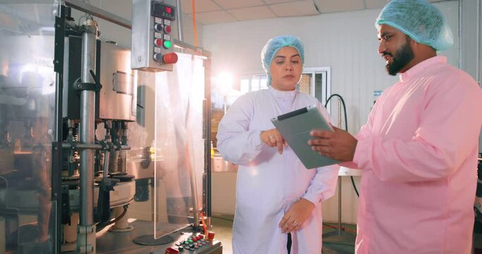 In an industrial water facility, two food scientist workers wearing hairnets and face masks converse while verifying quality control and taking notes on juice bottles. beverage production line inspect