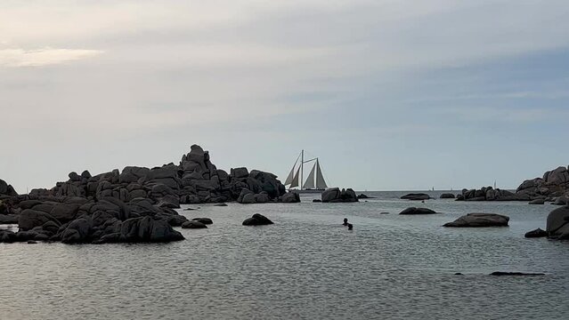 Two-masted sailing ship crosses the scene at Cala della Chiesa spot on Lavezzi island in Corsica with people bathing, France