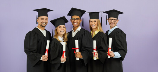 Happy diverse mixed race international university students in graduate hats and gowns showing diploma scrolls with red ribbons and smiling at camera standing on color background at graduation event