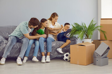 Family having fun together. Young family with children who are moving to new house take break, have fun and fool around. Mom, dad and sister tickle little boy sitting on sofa between cardboard boxes. - obrazy, fototapety, plakaty