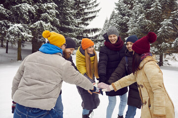 Group of cheerful friends having some outdoor fun among trees in snowy winter park. Team of happy young people in warm hats, coats and jackets standing in circle and putting their hands together