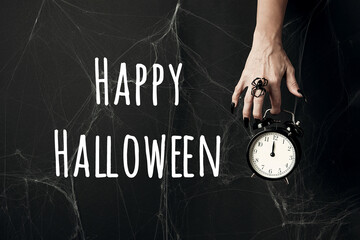 Halloween holiday greeting card. Happy Halloween text and hand with long black nails and spider ring holds alarm clock on black background with cobwebs