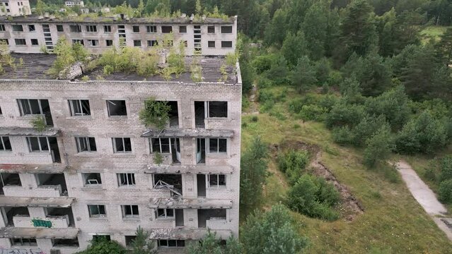 Irbene, Latvia - Fly along the front of an old udssr soviet buildings in the ghost town Irbene covered with plants and trees without windows - lost and forgotten in the middle of the forest bird view
