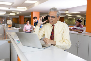 Worried senior businessman talking on mobile phone while using laptop at business desk