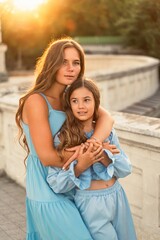 Portrait of mother and daughter in blue dresses with flowing long hair against the backdrop of sunset. The woman hugs and presses the girl to her. They are looking at the camera.