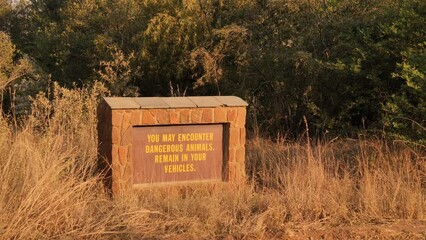 Stone built structure amid grass holds sign board in wildlife area. Yellow letters on faded maroon metal sign warn tourists to stay in vehicle due to dangerous animals