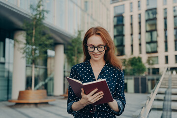 Outdoor shot of red haired woman makes notes in notepad makes plannings for working day