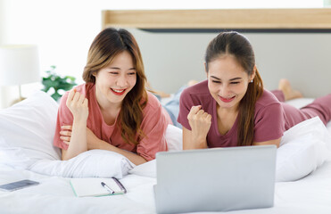 Millennial Asian young happy cheerful female teenager LGBTQ lesbian lover couple laying down together on bed and pillow learning studying online via laptop computer and writing short note on notebook