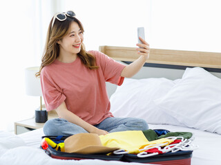 Millennial Asian young happy cheerful female teenager with sunglasses sitting smiling on cozy bed in bedroom at home holding smartphone taking selfie photo while packing clothes for holiday vacation