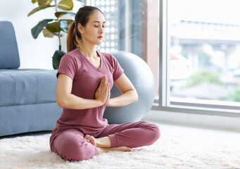 Millennial Asian young healthy female housewife sitting on carpet floor in living room doing hobby yoga exercising with lotus pose position meditating alone for good body care health and wellness