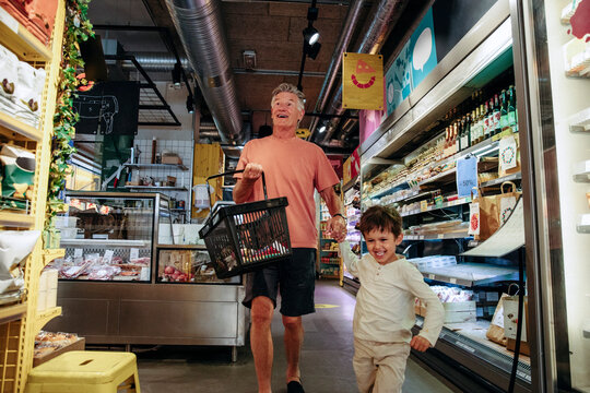 Happy grandson pulling grandfather holding basket while shopping at supermarket