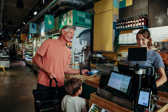 Happy senior man talking to cashier while standing with grandson at checkout counter in supermarket