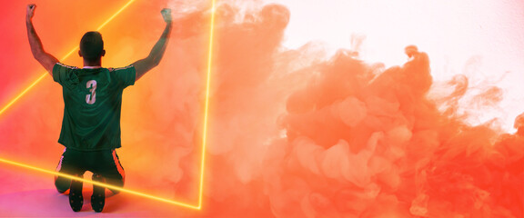 Rear view of caucasian player with arms raised kneeling over illuminated triangle and orange smoke