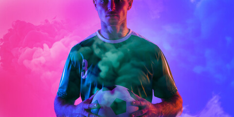 Male caucasian soccer player holding ball standing againsy smoky pink and blue background