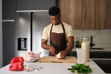 Fototapeta na wymiar Young black man cutting onion to prepare chicken recipe in a kitchen.