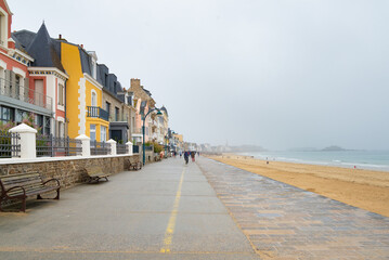 The pedestrian promenade of Saint Malo, Brittany, France, on a rainy day. Traditional buildings on the left. The shore with atlantic ocean on the right. White sky with copy-space on the background.