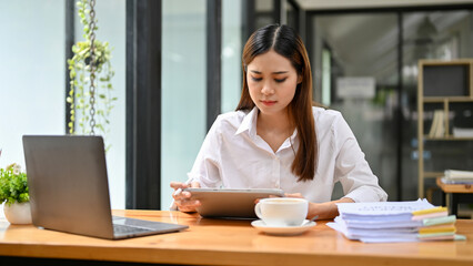 Fototapeta premium Beautiful Asian businesswoman concentrating on her work, using tablet, sitting at her desk.