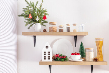wooden open kitchen shelves on a white textured wall with cans for loose products, a bouquet of spruce branches, balls and a cardboard white house.