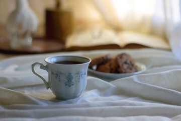 Cup of tea or coffee, plate of chocolate chip cookies, open book, reading glasses, candle and vase with flowers. Selective focus.