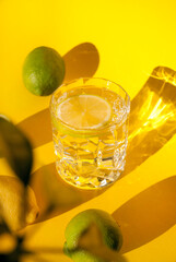 Glass of water with lemon slice with yellow and green lemons fruit on yellow background with shadow. Refreshing summer beverage. Summer cold drink cocktail