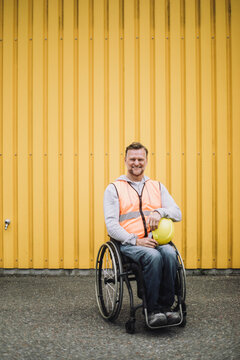 Happy construction worker sitting with hardhat in wheelchair against yellow metal wall